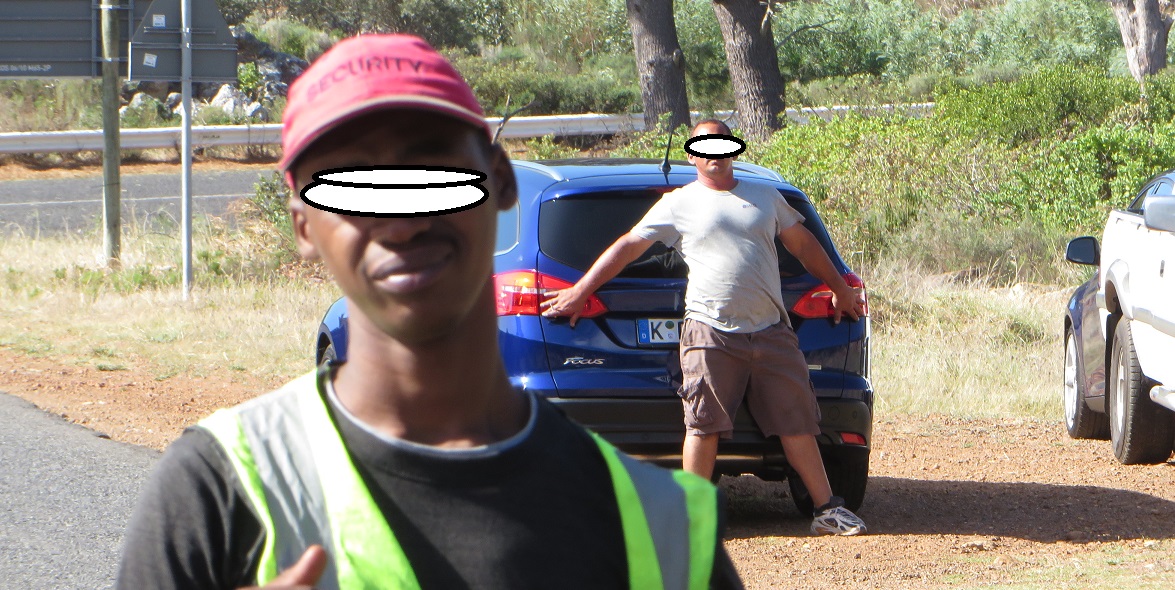 Ein Mann versucht den neuen Ford während des Drehs zum neuen Werbespots am Kap der Guten Hoffnung zu verdecken. Ein farbiger Mitarbeiter kommt auf den Fotografen drohend zu.
