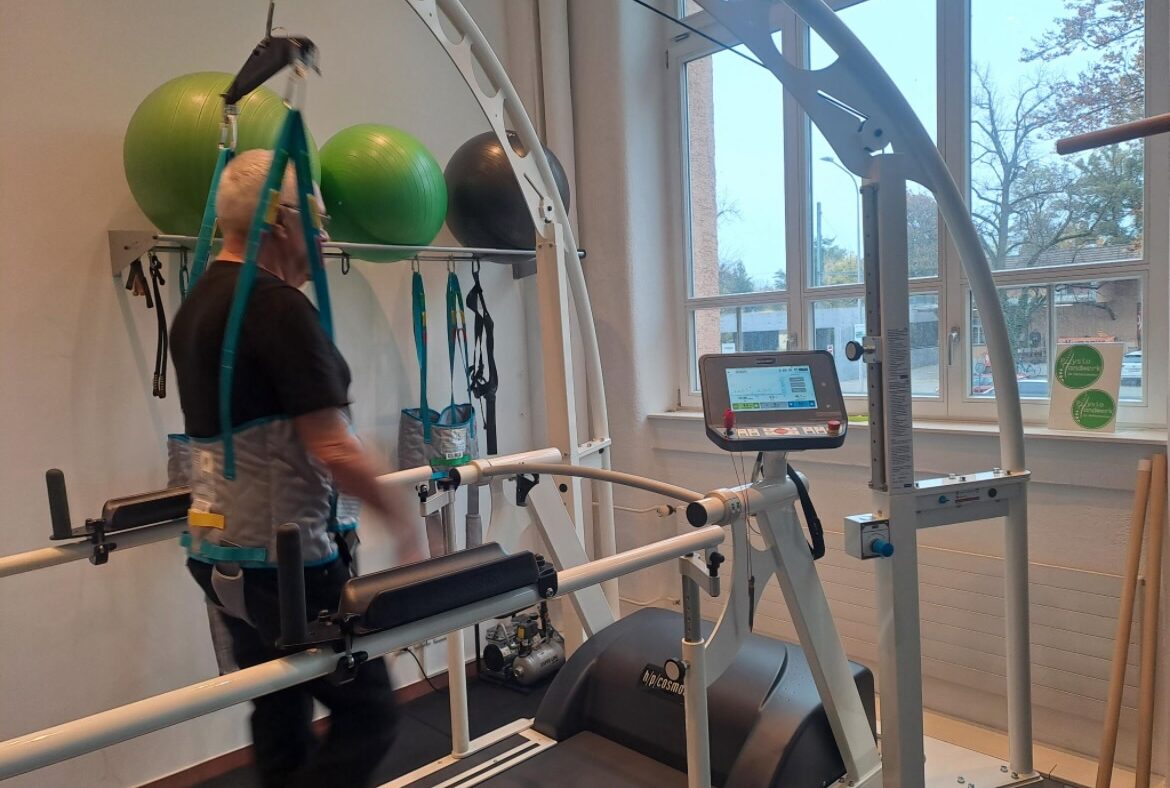 Ein Patient mit Sicherungsgurt geht unter Anleitung auf dem h/p cosmos Perturbationslaufband, während im Hintergrund Gymnastikbälle und Therapiehilfen bereitliegen. Fotoquelle: NETZ-TRENDS.de / Physiohandwerk – Physiotherapie Kreuzlingen, Sven Gruber am Bodensee, Schweiz