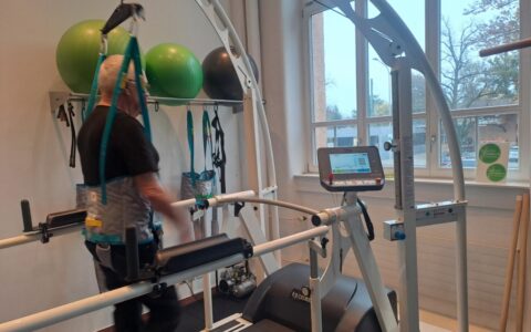Ein Patient mit Sicherungsgurt geht unter Anleitung auf dem h/p cosmos Perturbationslaufband, während im Hintergrund Gymnastikbälle und Therapiehilfen bereitliegen. Fotoquelle: NETZ-TRENDS.de / Physiohandwerk – Physiotherapie Kreuzlingen, Sven Gruber am Bodensee, Schweiz