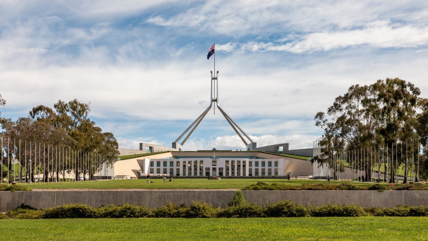 Das Parlamentsgebäude in Canberra mit der australischen Flagge auf dem Fahnenmast, davor eine Rasenfläche und Eukalyptusbäume