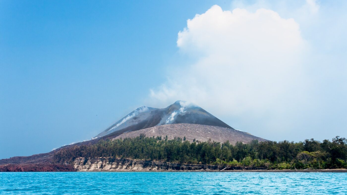Der Vulkan Anak Krakatau erhebt sich als kahler grauer Kegel ueber einer bewaldeten Insel in der Sundastrasse; an mehreren Stellen des Hangs steigt weisser Dampf auf.