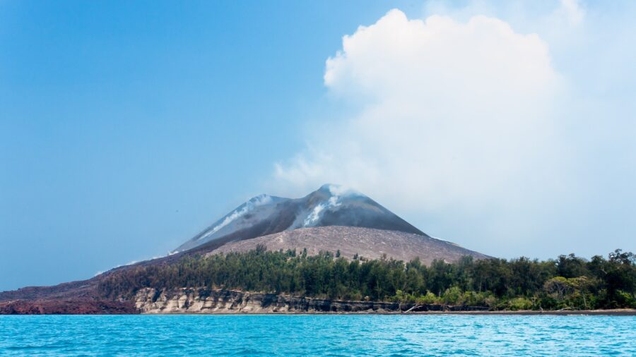 Der Vulkan Anak Krakatau erhebt sich als kahler grauer Kegel ueber einer bewaldeten Insel in der Sundastrasse; an mehreren Stellen des Hangs steigt weisser Dampf auf.