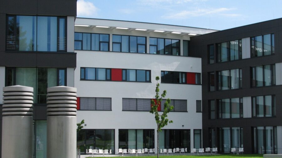 Modernes weisses Buerogebaeude mit dunklen Fensterrahmen und roten Farbakzenten, davor eine Rasenflaeche mit jungem Baum und weissen Stuehlen.
