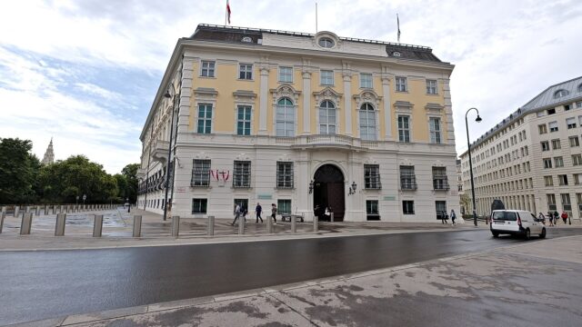 Das Bundeskanzleramt am Ballhausplatz in Wien, ein gelb-weisser Barockbau mit oesterreichischer Fahne auf dem Dach, davor Passanten und eine leere Strasse