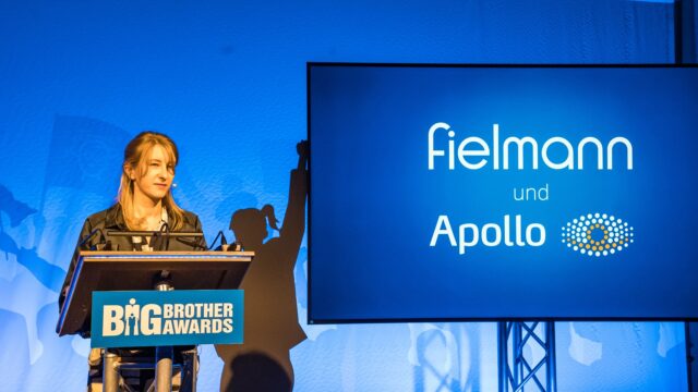Elisabeth Niekrenz am Rednerpult der BigBrotherAwards 2026, daneben eine Leinwand mit den Schriftzuegen Fielmann und Apollo