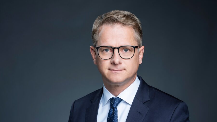 Porträtfoto von Bundesgesundheitsminister Carsten Linnemann im dunklen Anzug mit Brille vor grauem Hintergrund