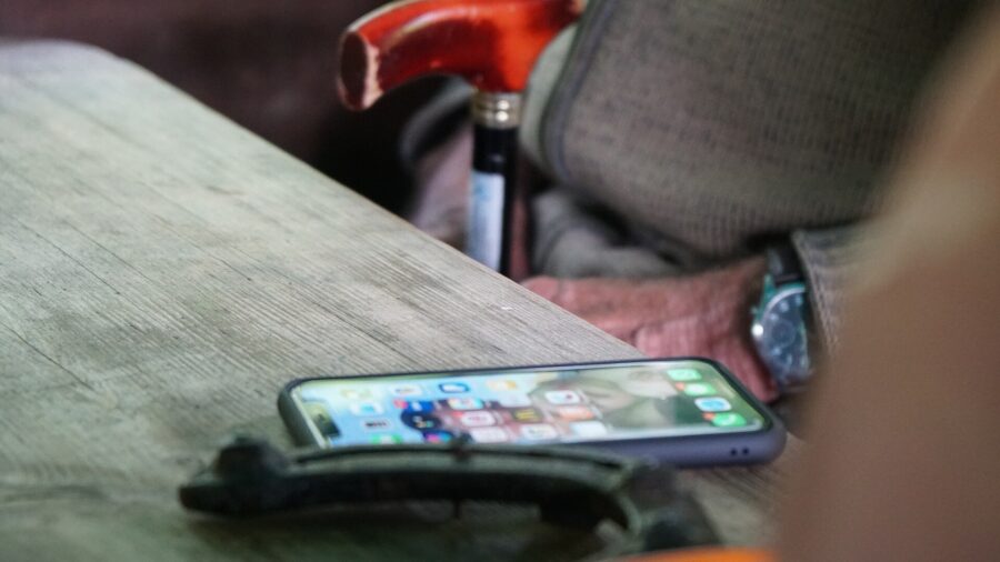 iPhone 11 mit eingeschaltetem Startbildschirm liegt auf der Kante eines Holztisches, im unscharfen Hintergrund ein Hammer und eine Hand
