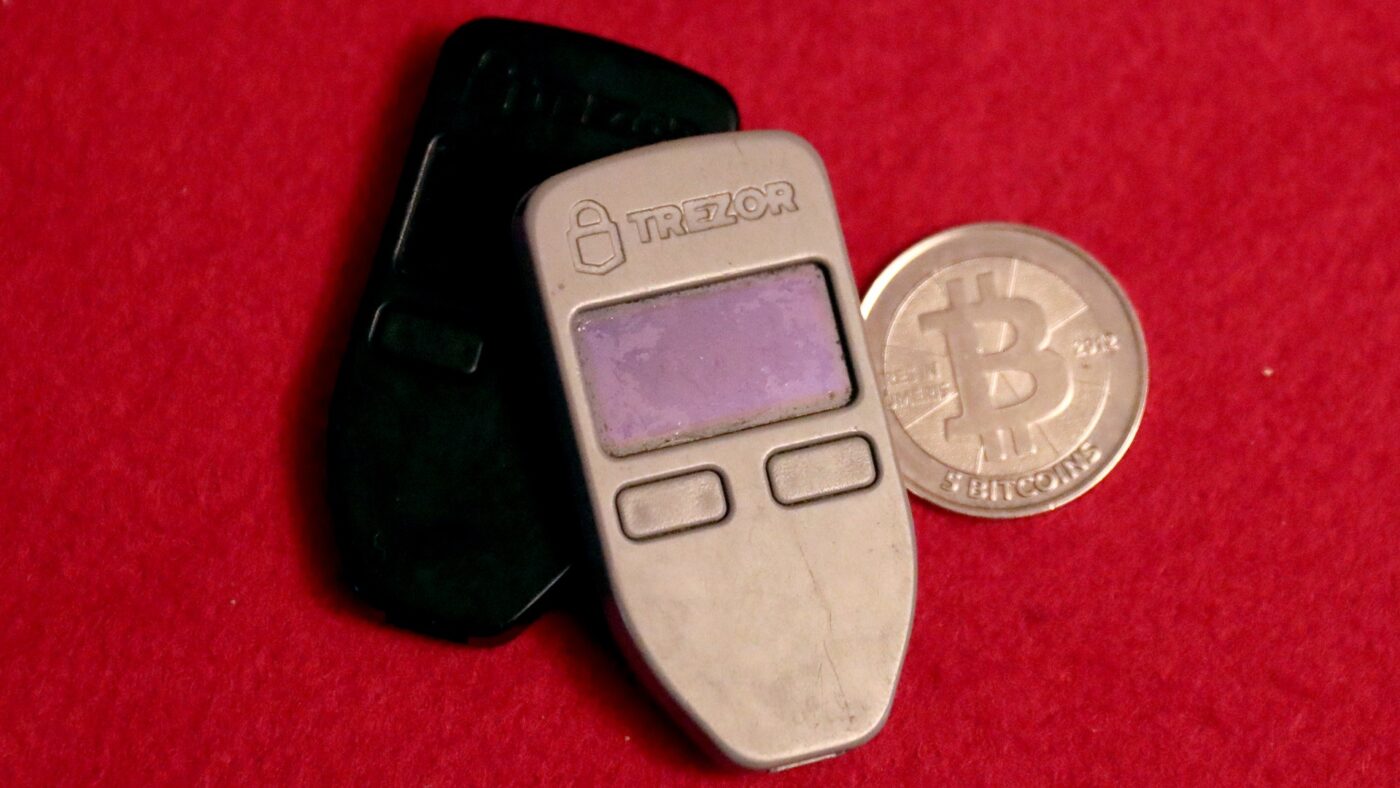 Zwei silberne und schwarze Hardware-Wallets des Herstellers Trezor liegen neben einer physischen Bitcoin-Muenze auf rotem Filzuntergrund.