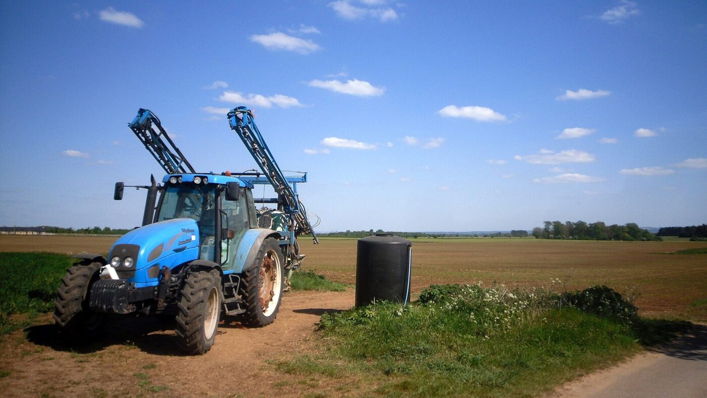 Blauer Traktor mit zusammengeklapptem Spritzgestaenge einer Feldspritze am Rand eines Ackers unter blauem Himmel