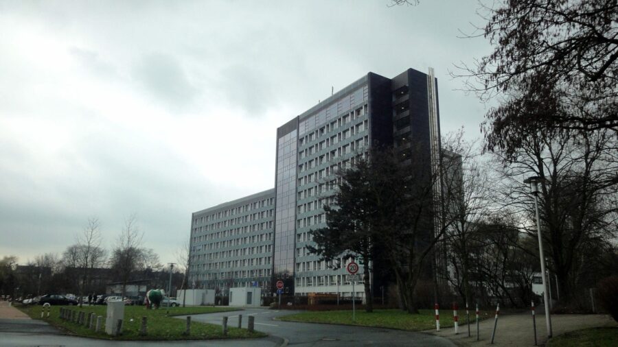 Das Hochhaus des Oberlandesgerichts Hamm bei bedecktem Himmel, davor eine Zufahrt mit Pollern, Wiese und kahlen Bäumen.