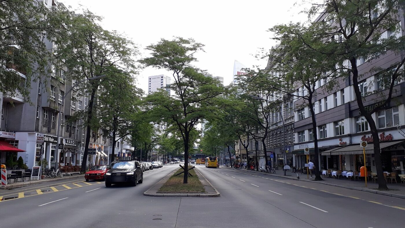 Blick über die mehrspurige Kantstraße in Berlin-Charlottenburg mit Mittelstreifen, Straßenbäumen, parkenden Autos und einem gelben Linienbus; im Hintergrund ein Hochhaus am Bahnhof Zoo.