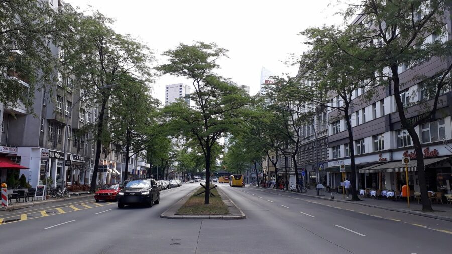 Blick über die mehrspurige Kantstraße in Berlin-Charlottenburg mit Mittelstreifen, Straßenbäumen, parkenden Autos und einem gelben Linienbus; im Hintergrund ein Hochhaus am Bahnhof Zoo.