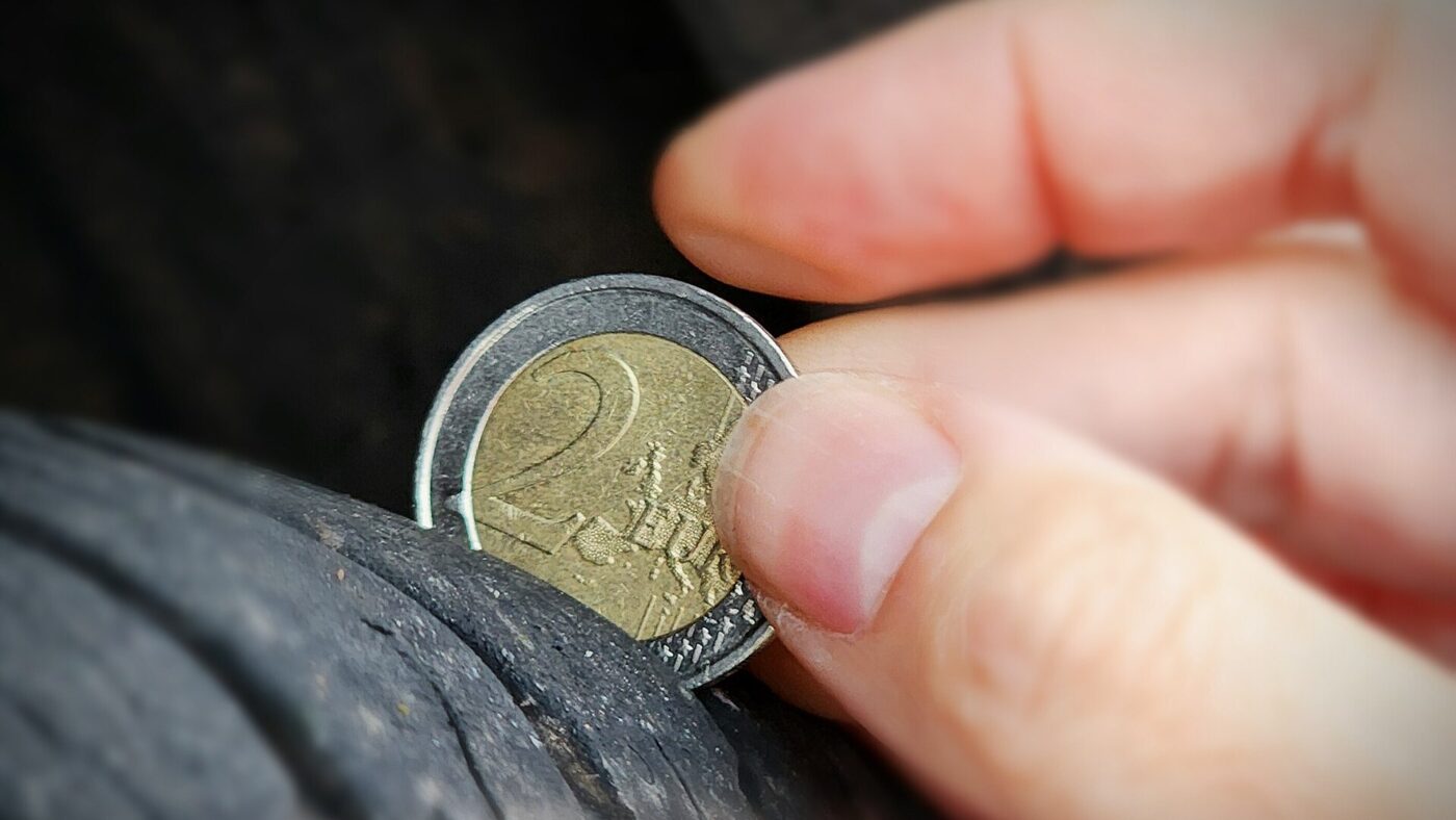 Nahaufnahme: Eine Zwei-Euro-Münze wird von einer Hand in eine Profilrille eines Autoreifens gesteckt, um die Restprofiltiefe zu prüfen.