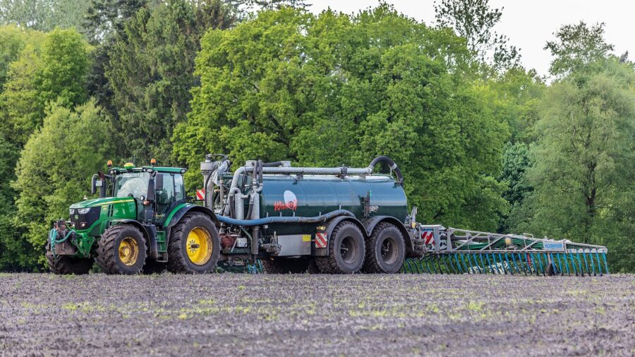 Ein gruener Traktor zieht ein dunkles Guellefass mit ausgeklapptem Schleppschlauchverteiler ueber einen frisch bestellten Acker, im Hintergrund eine Baumreihe.