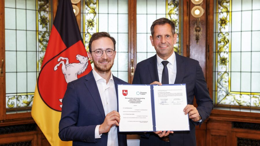 Zwei Männer in dunklen Anzügen halten in einem holzvertäfelten Saal vor der niedersächsischen Landesflagge eine aufgeschlagene Urkundenmappe mit dem Titel Memorandum of Understanding zum Aufbau einer HTS-Band-Produktion in Niedersachsen in die Kamera.