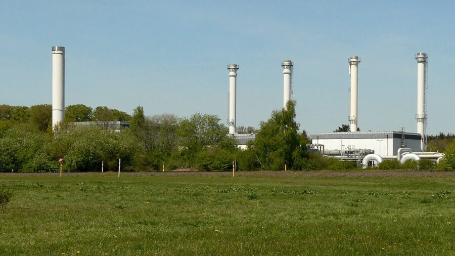 Obertägige Anlagen des Erdgasspeichers Rehden in Niedersachsen: fünf weiße Entlüftungskamine und flache Betriebsgebäude hinter einer Wiese und einer Baumreihe.