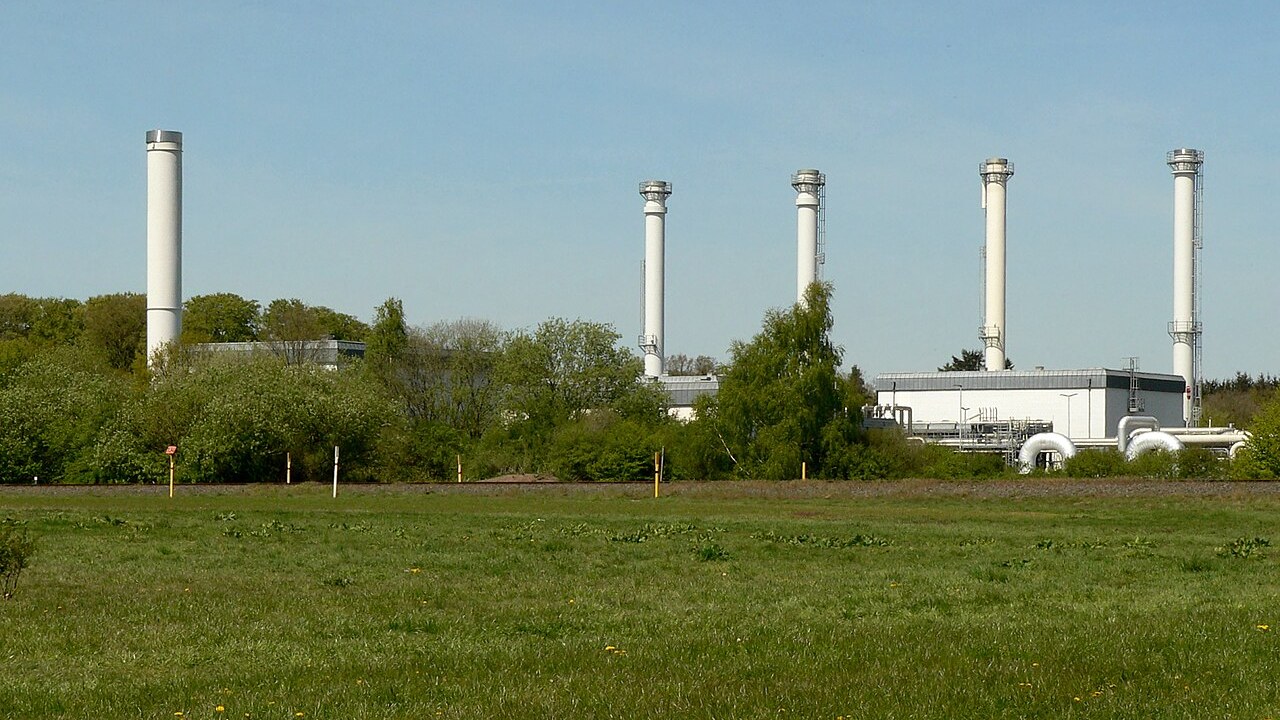 Obertägige Anlagen des Erdgasspeichers Rehden in Niedersachsen: fünf weiße Entlüftungskamine und flache Betriebsgebäude hinter einer Wiese und einer Baumreihe.