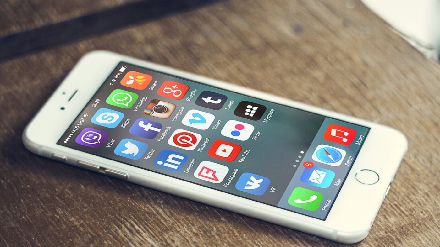 Weisses Smartphone auf einem Holztisch, auf dem Bildschirm die Symbole zahlreicher Apps sozialer Netzwerke