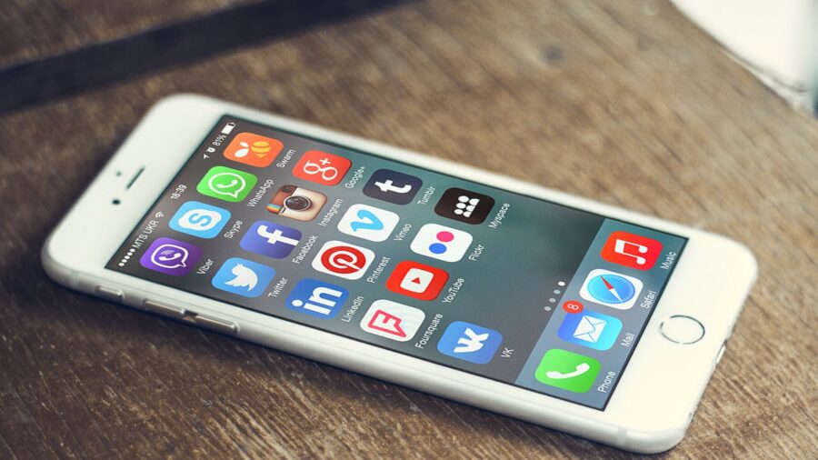 Weisses Smartphone auf einem Holztisch, auf dem Bildschirm die Symbole zahlreicher Apps sozialer Netzwerke