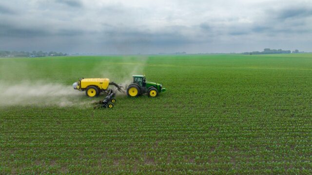 Traktor mit angehaengter Feldspritze faehrt ueber ein Feld mit jungen Pflanzen, Spruehnebel zieht seitlich ab