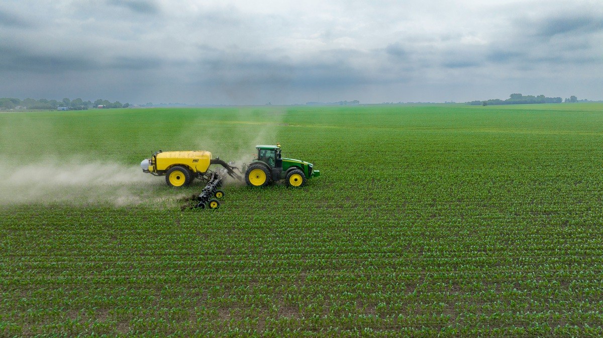 Traktor mit angehaengter Feldspritze faehrt ueber ein Feld mit jungen Pflanzen, Spruehnebel zieht seitlich ab