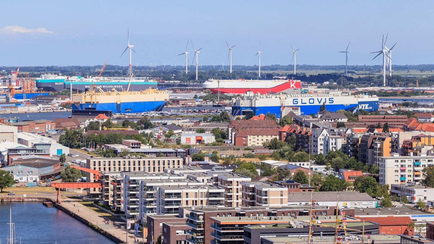 Blick über den Hafen von Bremerhaven mit mehreren Autotransportschiffen an den Kajen, Reihen abgestellter Neuwagen auf dem Terminal und Windenergieanlagen am Horizont
