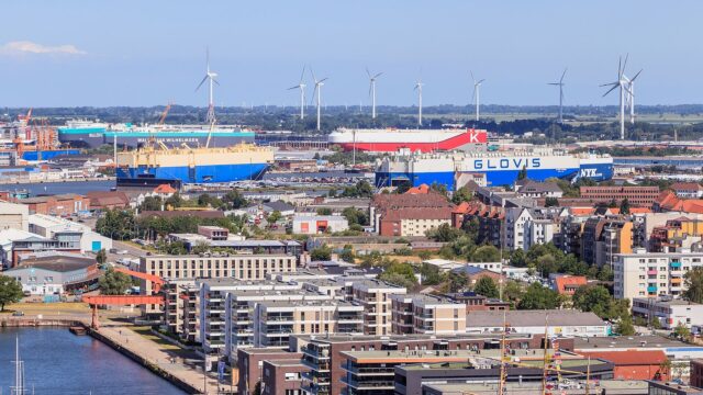 Blick über den Hafen von Bremerhaven mit mehreren Autotransportschiffen an den Kajen, Reihen abgestellter Neuwagen auf dem Terminal und Windenergieanlagen am Horizont