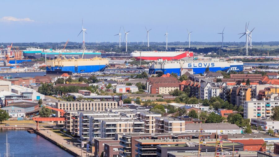 Blick über den Hafen von Bremerhaven mit mehreren Autotransportschiffen an den Kajen, Reihen abgestellter Neuwagen auf dem Terminal und Windenergieanlagen am Horizont
