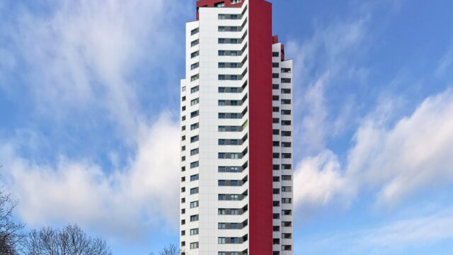 Weisses Wohnhochhaus mit roter Seitenfassade vor blauem Himmel mit Wolken, im Vordergrund kahle Baumkronen