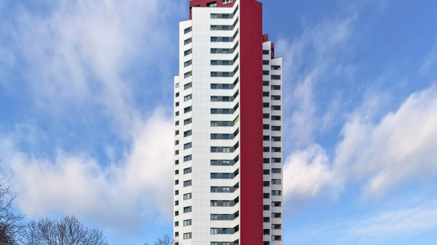 Weisses Wohnhochhaus mit roter Seitenfassade vor blauem Himmel mit Wolken, im Vordergrund kahle Baumkronen