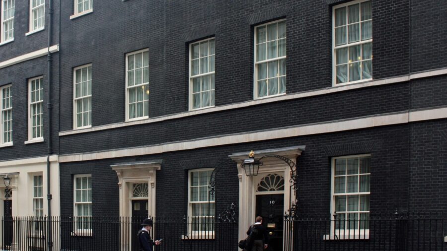 Dunkle Häuserfassade der Downing Street in London mit der schwarzen Eingangstür der Hausnummer 10, davor stehen ein Polizist und ein Passant.