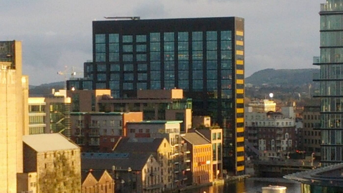 Hochhaus aus dunklem Glas mit gelb-schwarz gestreifter Seitenfassade im Dubliner Hafenviertel Grand Canal Dock, umgeben von niedrigeren Wohn- und Buerogebaeuden im Abendlicht