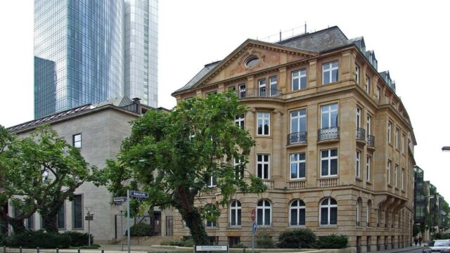 Eckgebäude der Bundesbank-Hauptverwaltung in Frankfurt am Main mit Hinweisschild, dahinter zwei Hochhäuser und Straßenbäume.