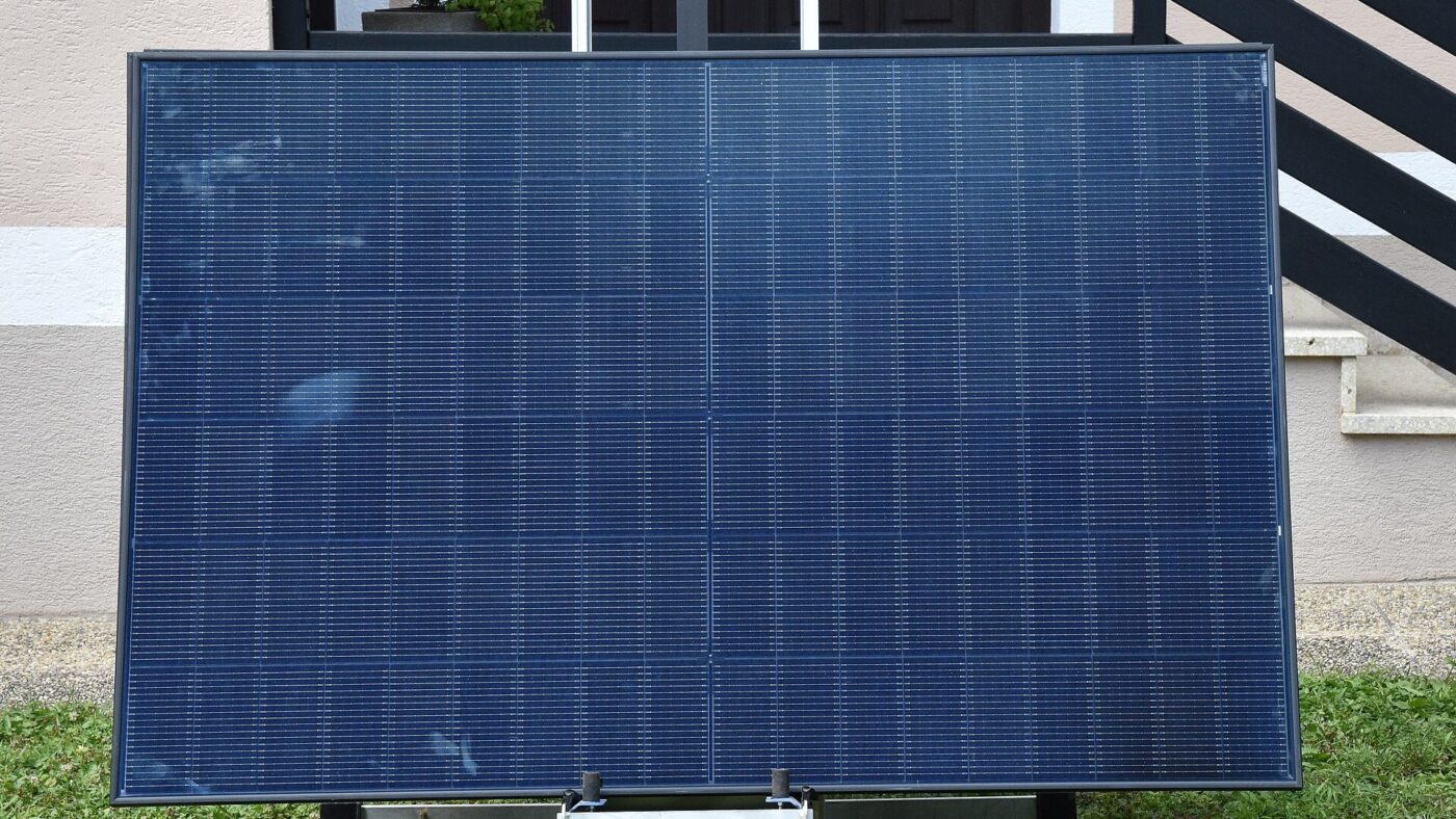 Ein Photovoltaikmodul steht schraeg aufgestellt auf einer Rasenflaeche vor der Fassade eines Wohnhauses.