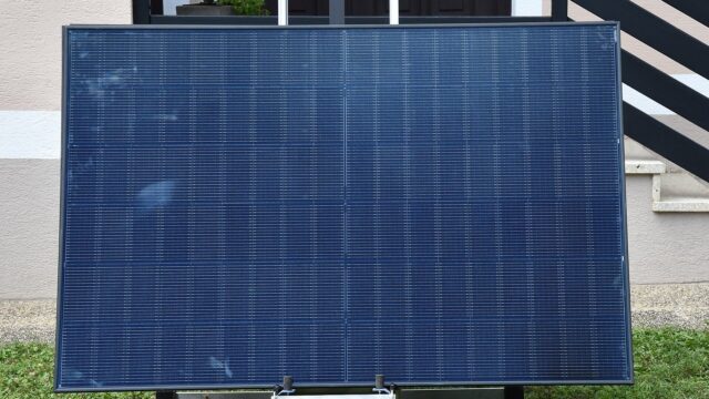 Ein Photovoltaikmodul steht schraeg aufgestellt auf einer Rasenflaeche vor der Fassade eines Wohnhauses.