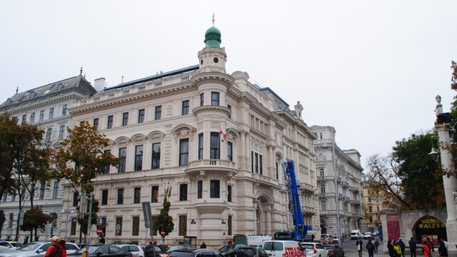 Gründerzeit-Eckhaus mit grüner Turmkuppel an einer Straßenkreuzung in der Wiener Innenstadt, davor parkende Autos, Bäume und Passanten