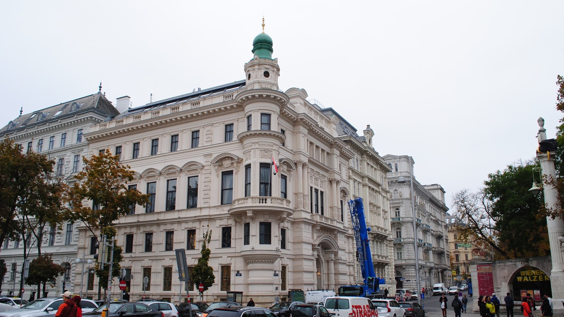 Gründerzeit-Eckhaus mit grüner Turmkuppel an einer Straßenkreuzung in der Wiener Innenstadt, davor parkende Autos, Bäume und Passanten