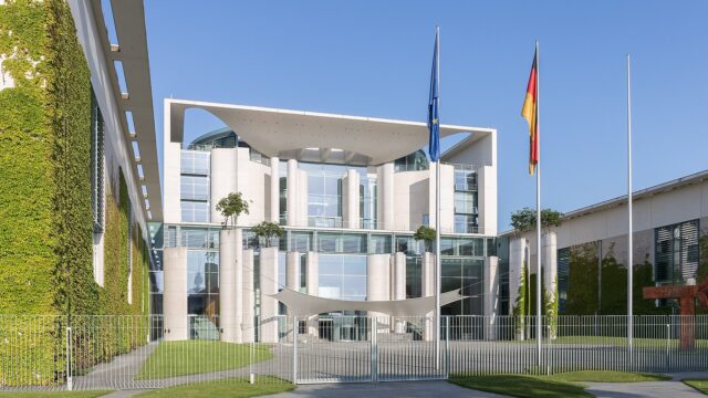 Das Bundeskanzleramt in Berlin bei Sonnenschein, davor die Flaggen Deutschlands und der Europaeischen Union.