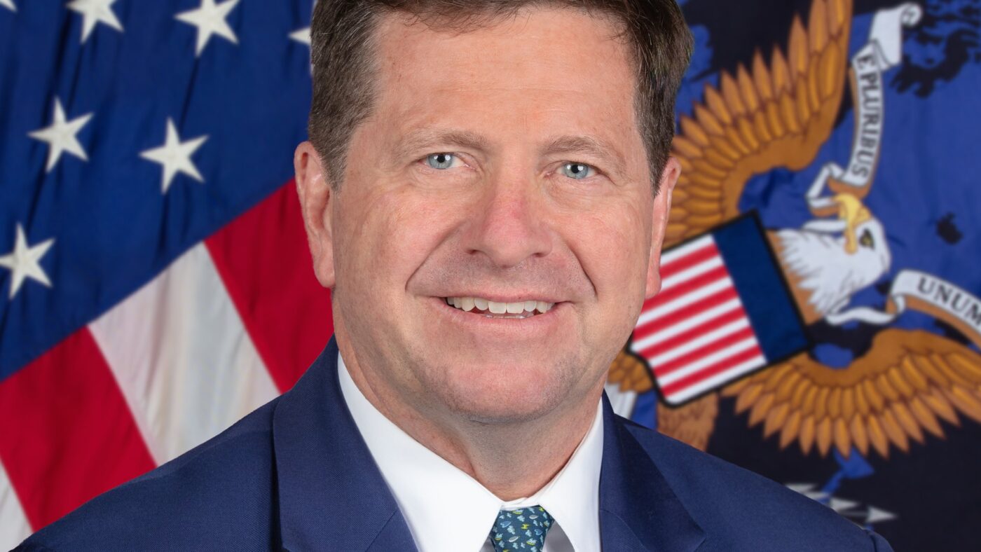 Porträtaufnahme von Jay Clayton im dunklen Anzug vor der US-Flagge und dem Dienstsiegel der US-Nachrichtendienste