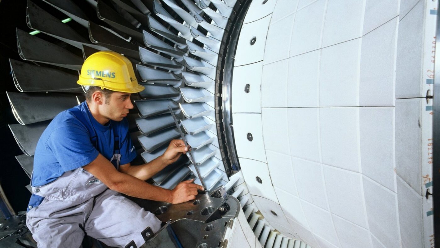 Ein Beschäftigter mit Schutzhelm montiert Laufschaufeln am Rotor einer großen Gasturbine in einer Werkshalle.