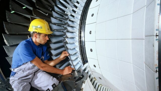 Ein Beschäftigter mit Schutzhelm montiert Laufschaufeln am Rotor einer großen Gasturbine in einer Werkshalle.