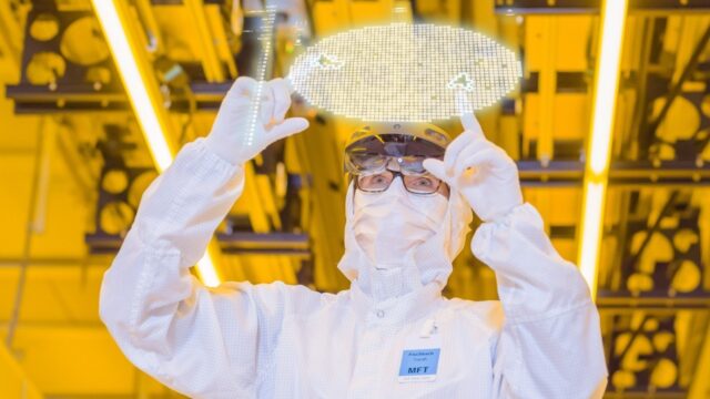 Person in weisser Reinraumkleidung haelt unter gelbem Licht einen runden Halbleiter-Wafer gegen die Beleuchtung in einer Chipfabrik