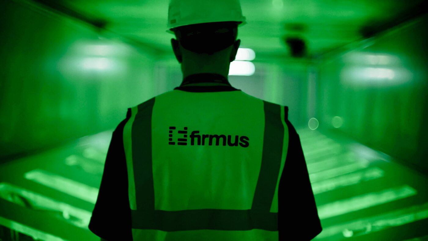 Person von hinten in einer Warnweste mit Firmus-Logo und weißem Schutzhelm in einem grün ausgeleuchteten Rechenzentrumsgang