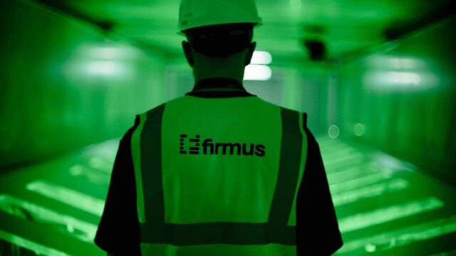 Person von hinten in einer Warnweste mit Firmus-Logo und weißem Schutzhelm in einem grün ausgeleuchteten Rechenzentrumsgang