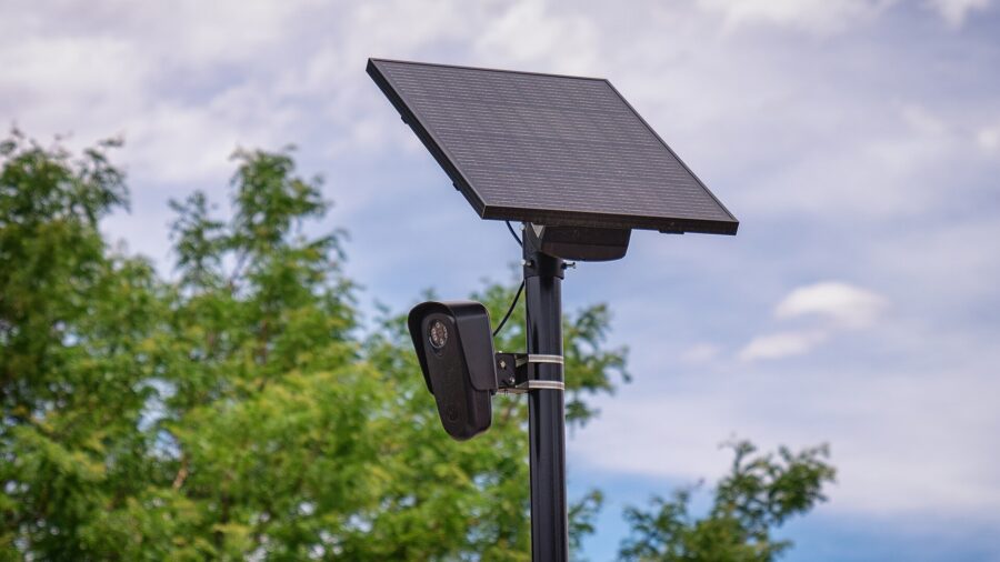 Schwarze Überwachungskamera mit Solarmodul an einem Mast vor Bäumen und bewölktem Himmel