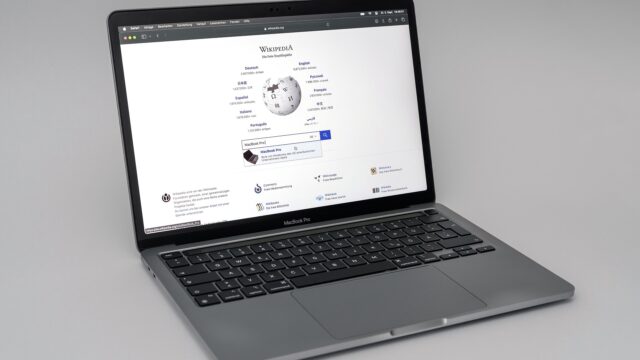 Aufgeklapptes MacBook Pro mit deutscher Tastatur auf hellgrauem Untergrund, auf dem Bildschirm die Startseite von Wikipedia.