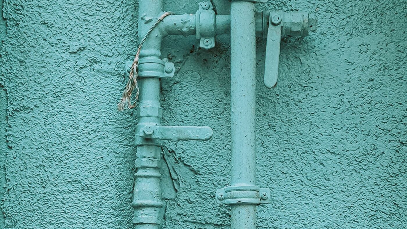 Mit türkiser Farbe gestrichene Gasrohre mit Absperrhähnen an einer verputzten Hauswand, Symbolbild für Gasnetzentgelte