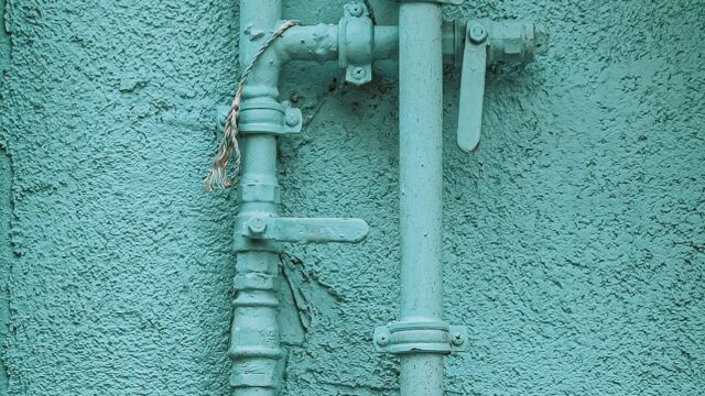 Mit türkiser Farbe gestrichene Gasrohre mit Absperrhähnen an einer verputzten Hauswand, Symbolbild für Gasnetzentgelte