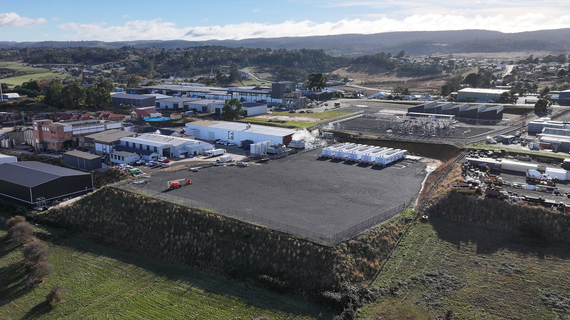 Luftaufnahme eines eingezäunten, asphaltierten Areals in einem Gewerbegebiet in Tasmanien; am Rand stehen mehrere weiße Containereinheiten, dahinter eine Umspannanlage und Hallen, im Hintergrund Hügel.