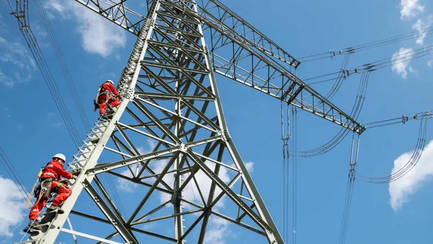 Zwei Monteure arbeiten an einem Hochspannungsmast beim Freileitungsbau vor blauem Himmel, Symbolbild zu den steigenden Strom-Umlagen 2027.